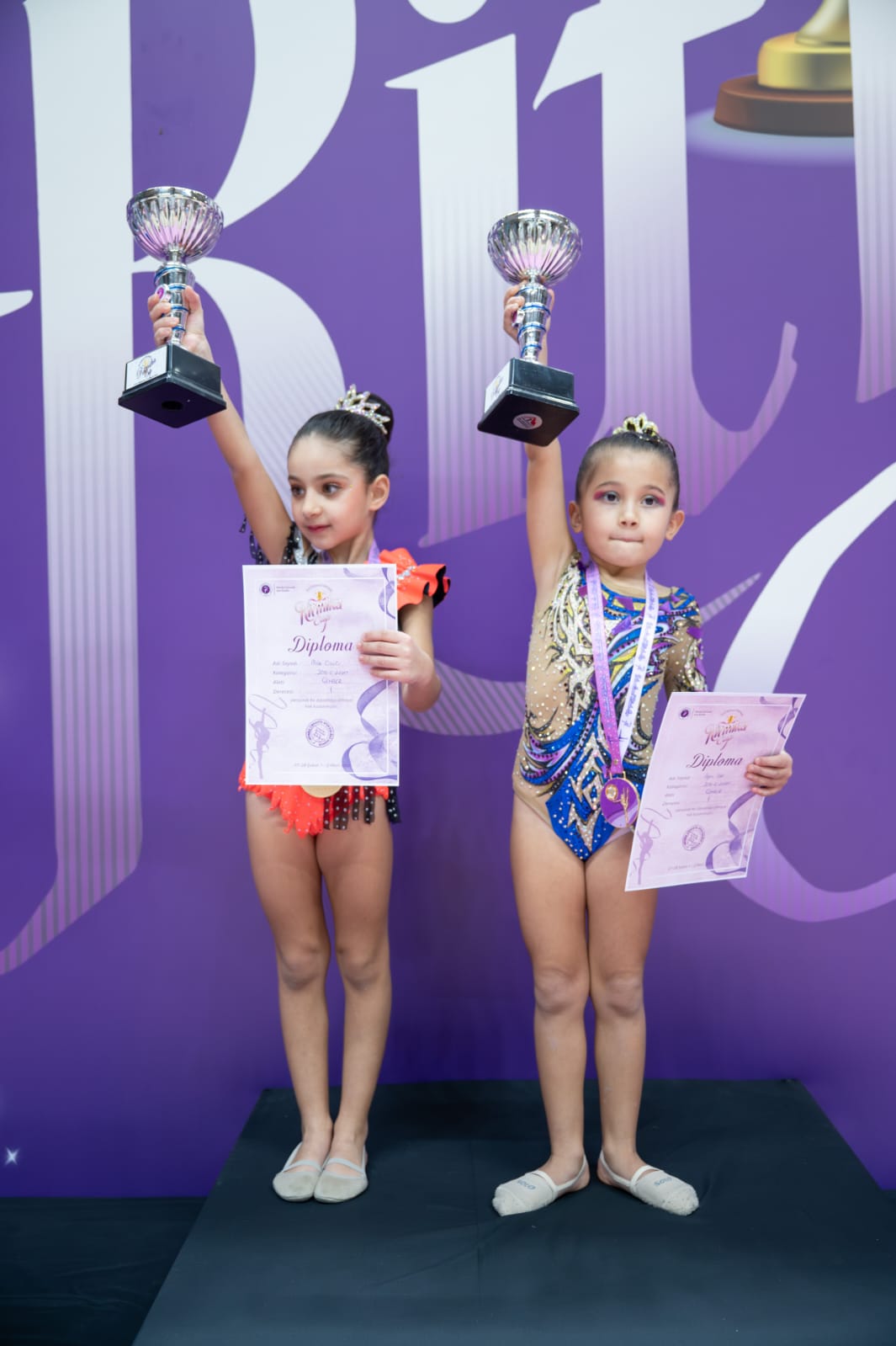 İki kız çocuğu, bir yarışmada ödüllerini ve diplomalarını tutarak gülümsüyor. Arka planda mor bir fon var.